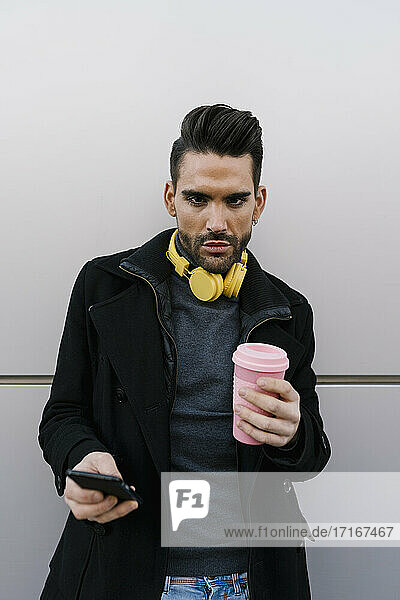 Handsome man with reusable cup holding smart phone against wall