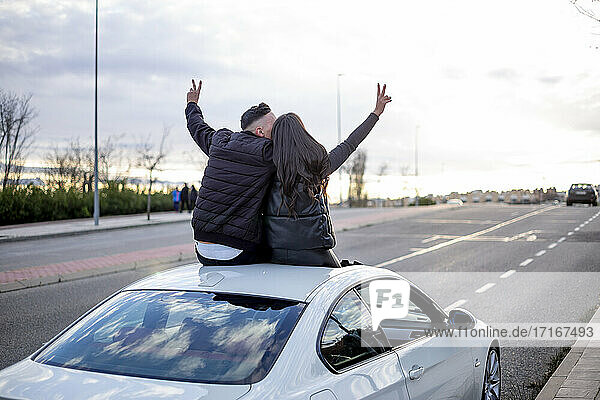 Unbeschwertes Paar gestikuliert Friedenszeichen  während es auf dem Autodach sitzt