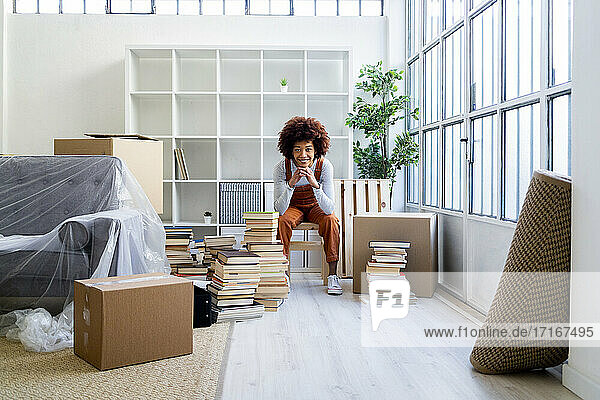 Lächelnde Afro-Frau mit Hand am Kinn  die in ihrem neuen Zuhause am Bücherregal sitzt