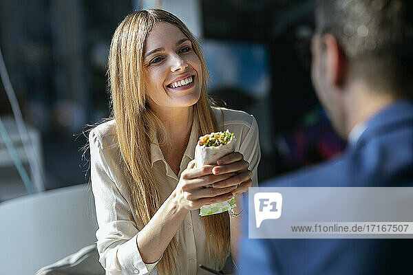 Geschäftsfrau mit Essen in der Hand und Blick auf einen in der Cafeteria sitzenden Kollegen