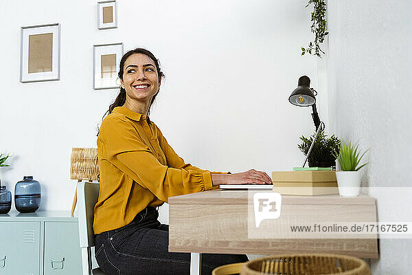 Smiling woman looking away while sitting with laptop by table at home