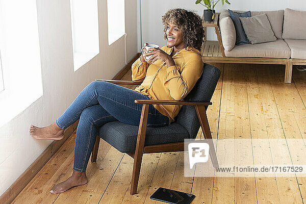 Lächelnde Frau trinkt Tee  während sie zu Hause auf einem Sessel sitzt