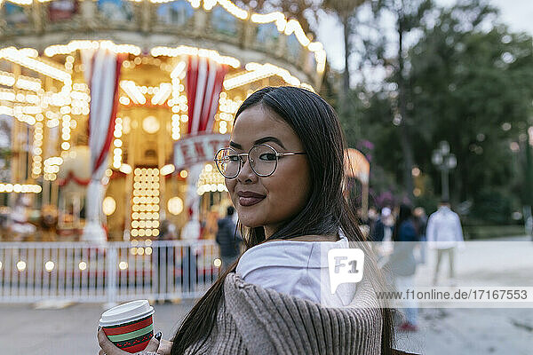 Beautiful young woman looking over shoulder while standing against illuminated carousel
