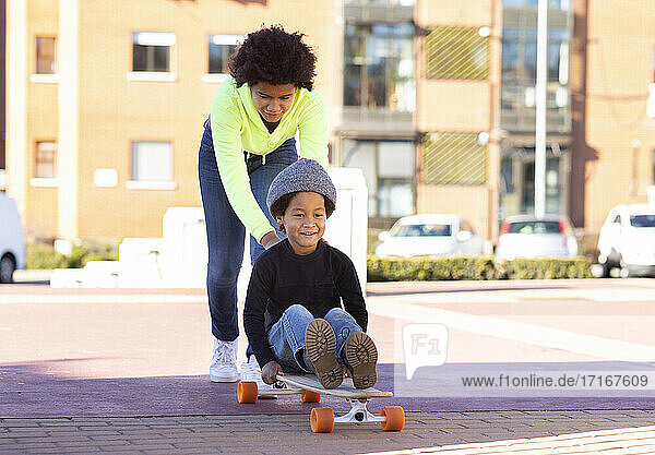 Schwester schiebt Bruder auf Skateboard sitzend beim Spielen auf dem Fußweg