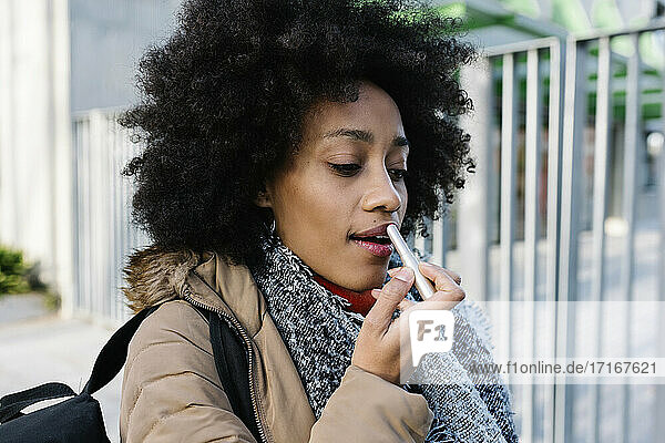 Afro young woman applying lipstick while standing on street