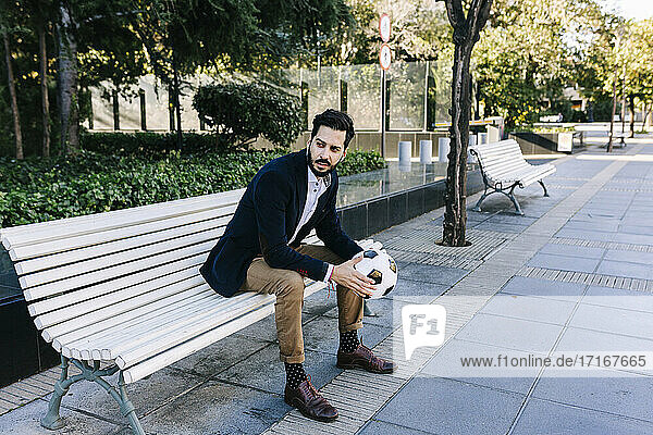 Businessman with soccer ball looking away while sitting on bench
