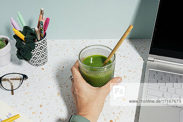 Woman's hand holding healthy green juice on desk at home