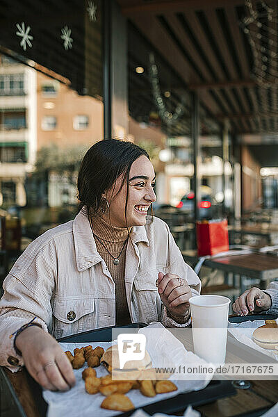 Cheerful teenage girl looking at friend while having fast food at outdoor restaurant