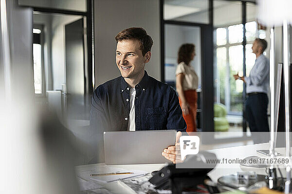Lächelnder Geschäftsmann  der wegschaut  während er mit Kollegen im Hintergrund in einem Großraumbüro sitzt