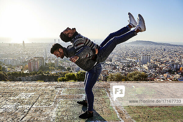 Verspielter Mann  der seinen Freund auf dem Rücken trägt  während er am Aussichtspunkt steht  Bunkers del Carmel  Barcelona  Spanien