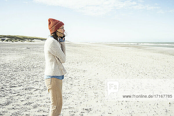 Porträt einer Frau mit Strickmütze  die am Strand mit ihrem Smartphone telefoniert