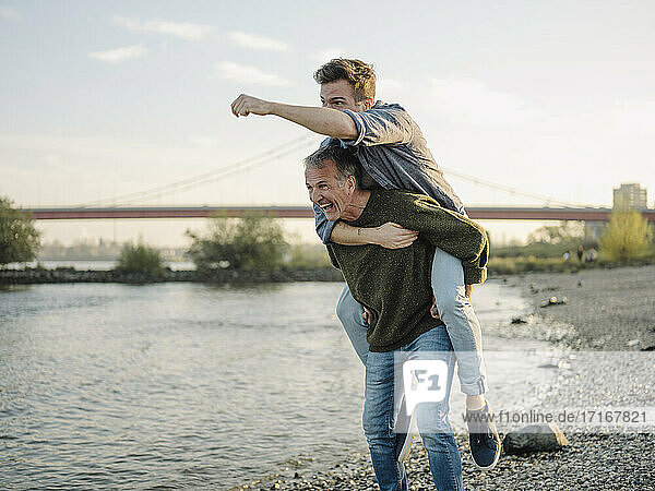 Happy father giving piggyback ride to son at riverbank against sky