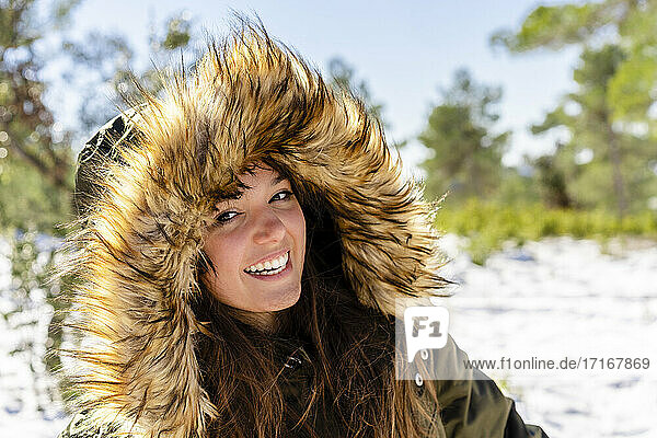Lächelnde junge Frau mit Pelzkapuzen-Wintermantel  die im Wald steht und starrt