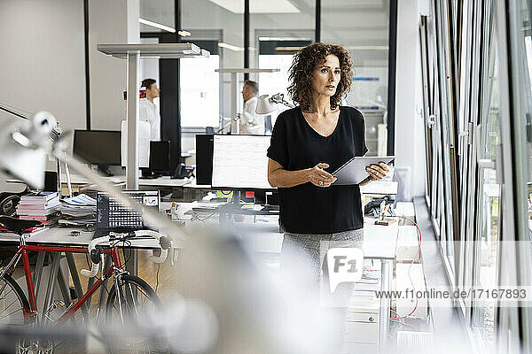 Geschäftsfrau hält digitales Tablet  während sie mit einem Kollegen im Hintergrund in einem Großraumbüro steht