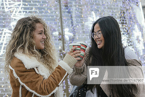Beautiful female friends toasting coffee cups against illuminated Christmas lights