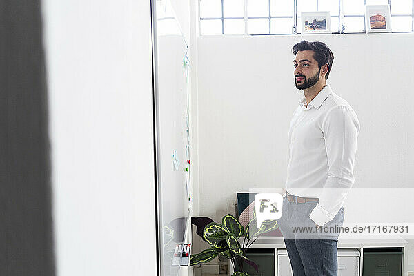 Smiling male entrepreneur with hands in pockets looking at whiteboard in office