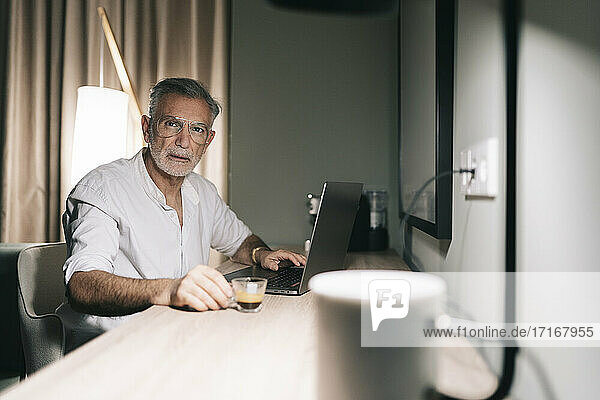Mature man holding coffee cup while sitting at desk with laptop in hotel room