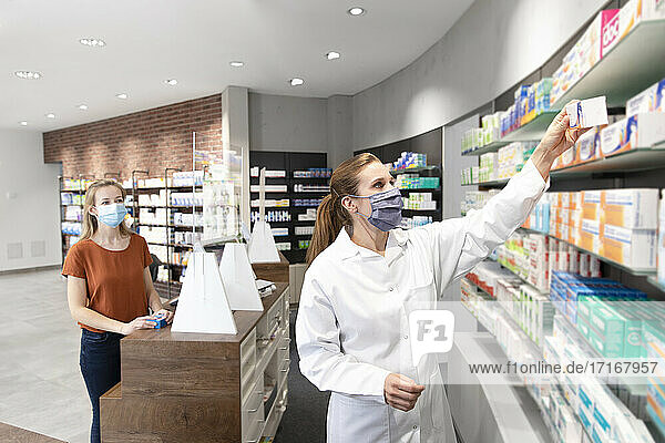 Customer standing at checkout looking at female pharmacist taking medicine from shelf in store
