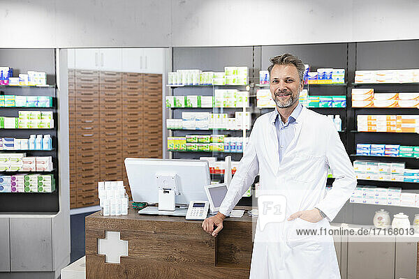 Mature male pharmacist with hand in pocket standing at pharmacy checkout