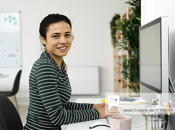 Glückliche weibliche Fachkraft sitzt auf dem Schreibtisch vor dem Computer am Arbeitsplatz