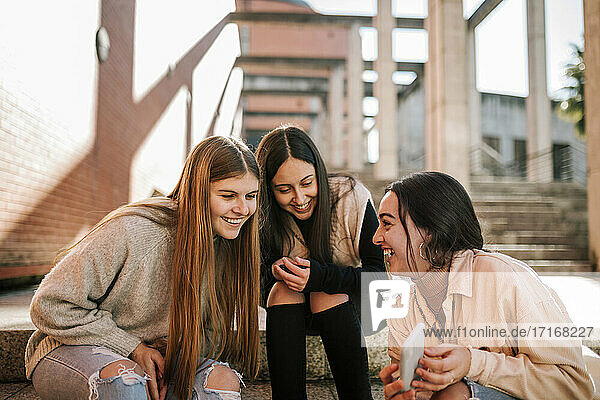 Fröhliches Teenager-Mädchen zeigt Freunden ihr Smartphone  während sie auf einer Treppe in der Stadt sitzt