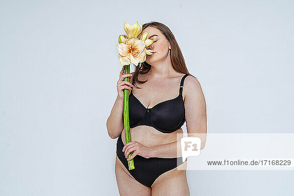 Junge Frau im schwarzen Bikini riecht an Blumen vor weißem Hintergrund
