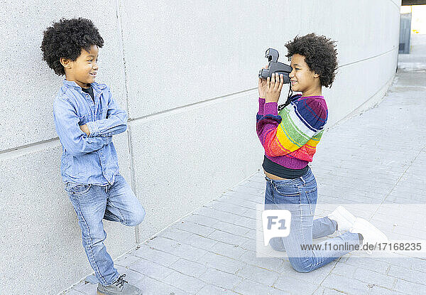 Sister taking photo of brother through camera while kneeling on footpath