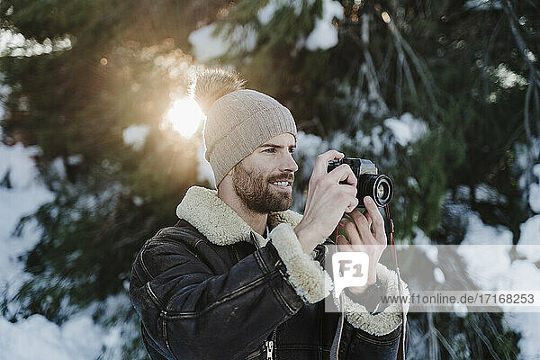 Handsome man photographing through camera against trees during winter