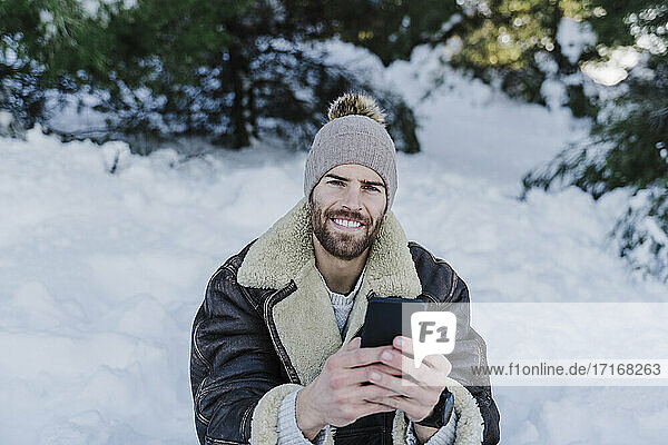 Lächelnder junger Mann mit Handy in der Hand  der im Schnee sitzt