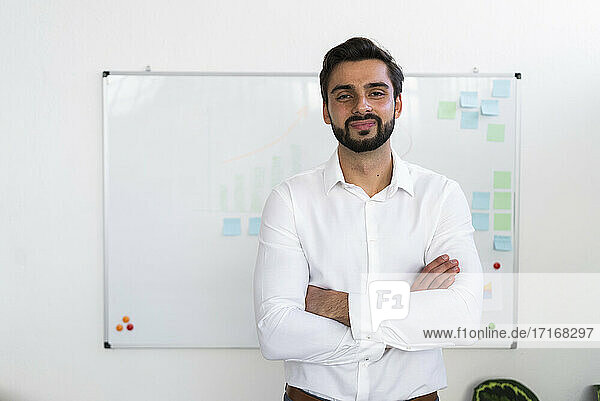 Lächelnder männlicher Unternehmer mit verschränkten Armen vor einem Whiteboard im Büro stehend