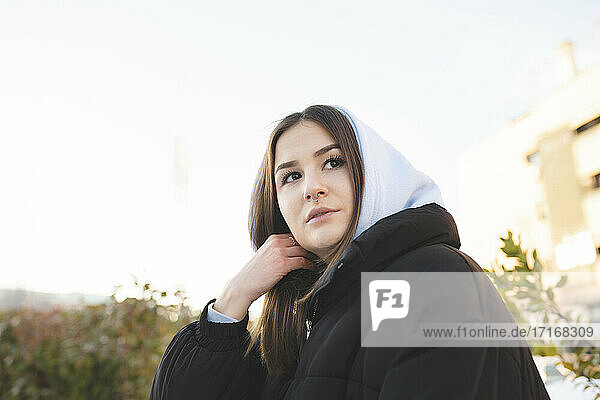 Young woman wearing hood clothing looking away while standing against sky