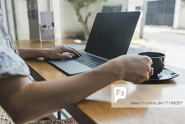 Hände einer Geschäftsfrau mit Kaffeetasse auf dem Tisch  die über einem Laptop in einem Café arbeitet
