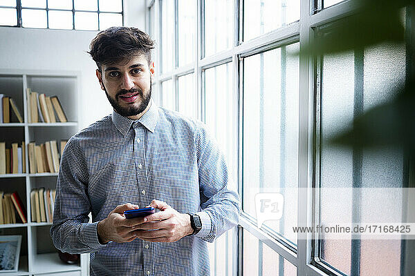 Porträt eines jungen Geschäftsmannes mit Telefon in der Hand  stehend am Fenster in einem kreativen Büro