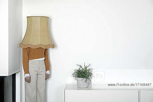 Mature woman with lamp shade on head standing against white wall