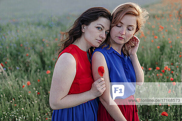 Woman holding flower while leaning on female friend's shoulder in poppy field
