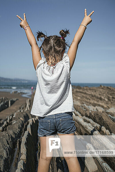 Girl with arms raised gesturing peace sign while standing on flysch against sky