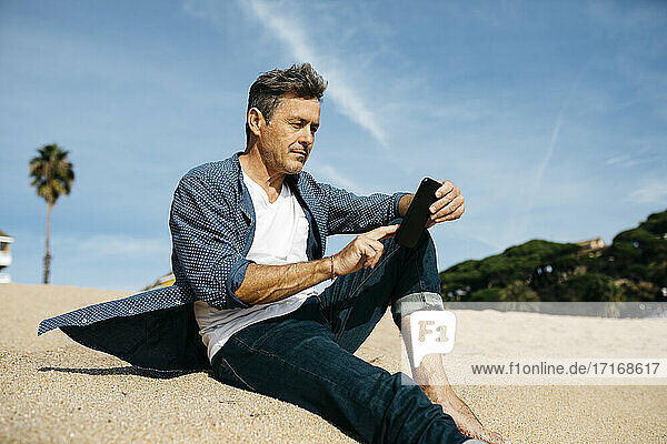 Älterer Mann  der ein Mobiltelefon benutzt  während er an einem sonnigen Tag am Strand sitzt