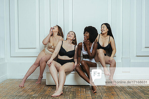 Multi-ethnische Gruppe von weiblichen Modellen in Dessous  die lachend an einer weißen Wand sitzen