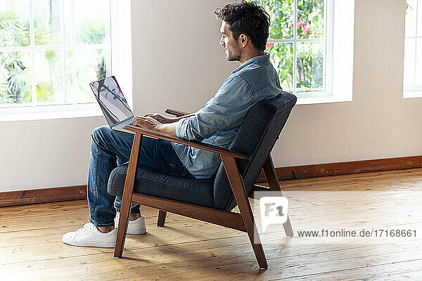 Mittlerer erwachsener Mann arbeitet am Laptop  während er zu Hause auf einem Sessel sitzt