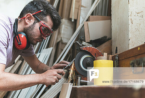 Concentrated male carpenter sharpening work tool on grinder in workshop
