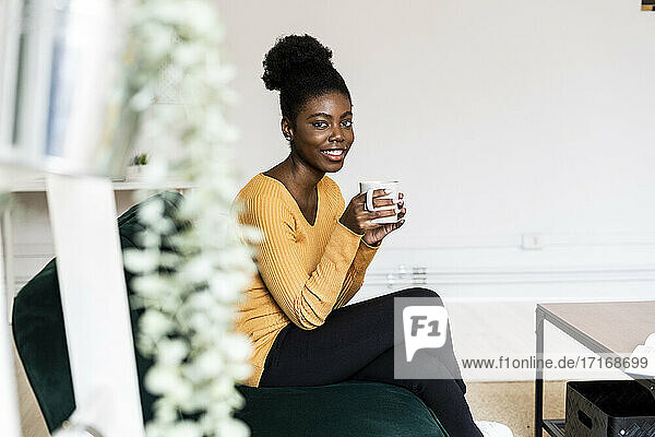 Smiling woman holding coffee cup while sitting on sofa at home