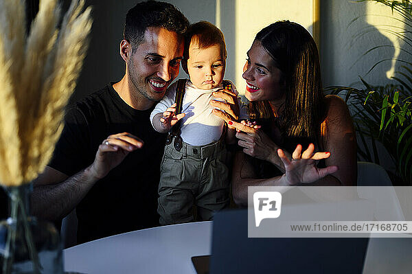 Parents with baby boy waving hand to video call on laptop while sitting at home