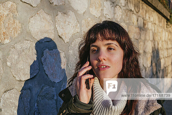 Nachdenkliche junge Frau  die bei Sonnenuntergang an einer Steinmauer mit ihrem Smartphone telefoniert
