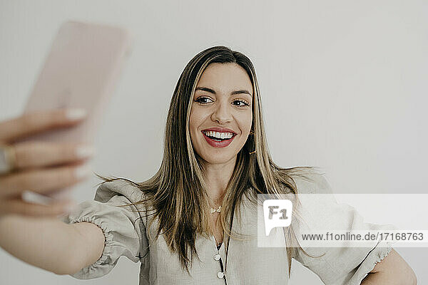 Smiling woman taking selfie through mobile phone against white background