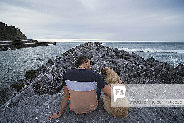 Man with arm around dog while sitting on rock against sky