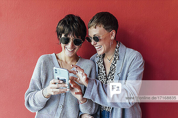 Glückliche Freunde  die ein Mobiltelefon benutzen  während sie vor einer roten Wand stehen