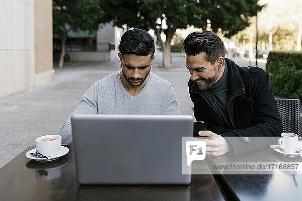 Male friends with laptop having coffee while sitting at side walk cafe in city