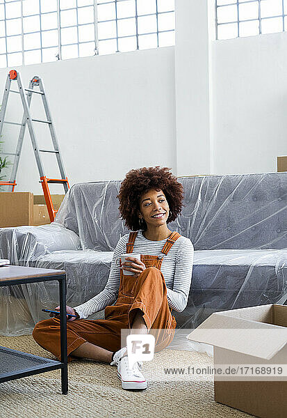 Afro-Frau mit Smartphone träumt  während sie eine Kaffeetasse in ihrer neuen Wohnung hält