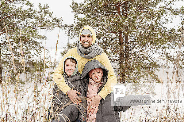 Vater  der seinen Sohn und seine Tochter umarmt  während er im Winter gegen Bäume im Wald steht