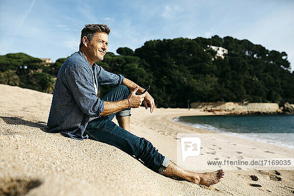 Lächelnder Mann mit Smartphone sitzt am Strand gegen Hügel während sonnigen Tag